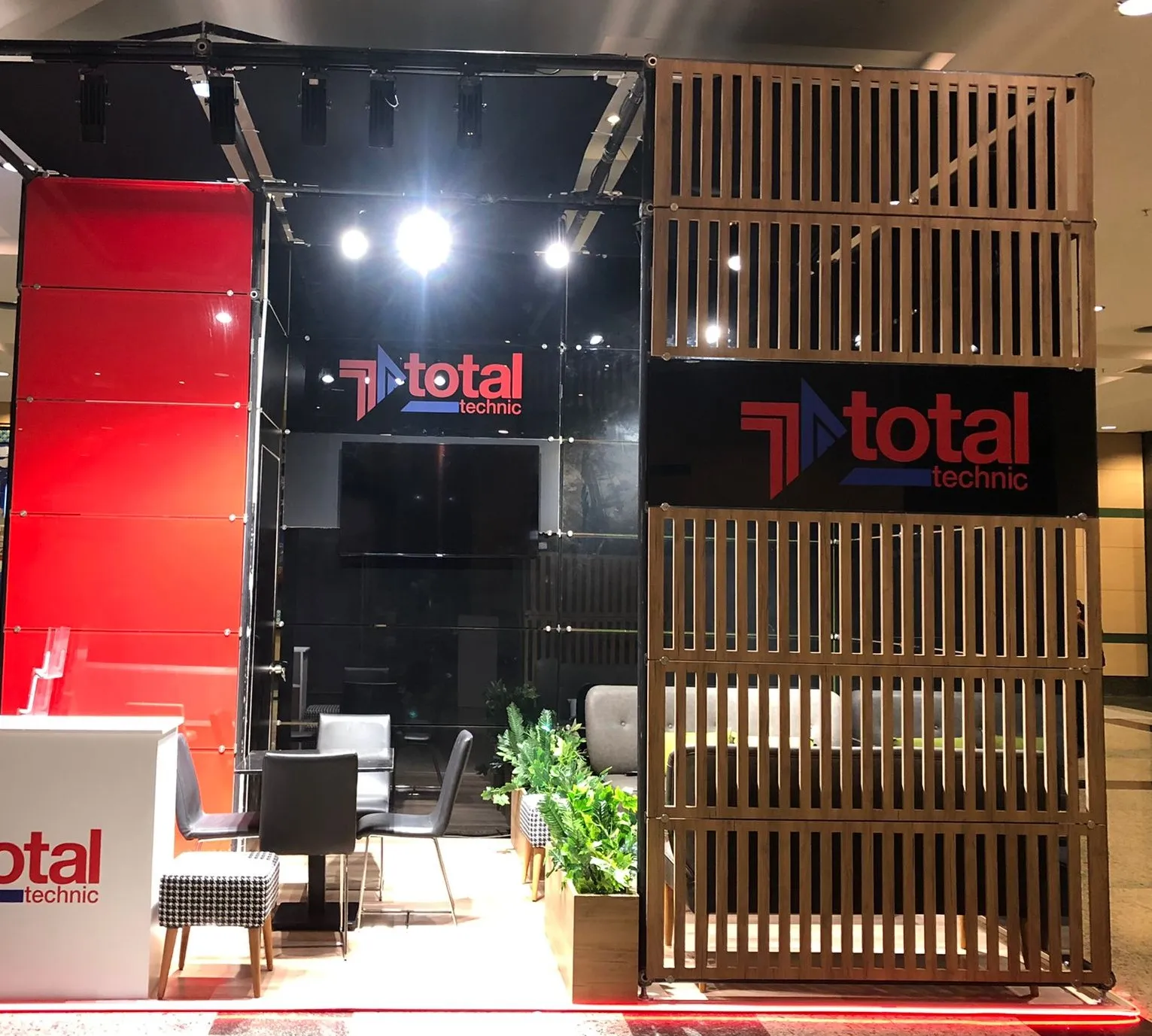 Total Technic için üretilmiş ahşap ve cam kombinasyonlu fuar standı; ışıklı alınlık, karşılama bankosu ve oturma alanı görünüyor.